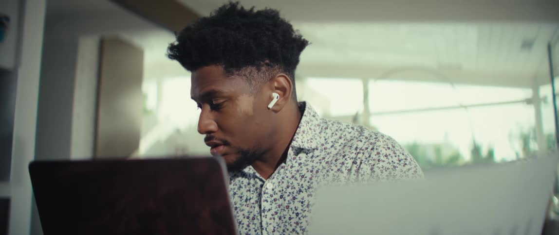 A man wearing a patterned shirt and wireless earbuds sits at a desk while looking at a document and using a laptop in a bright modern home office