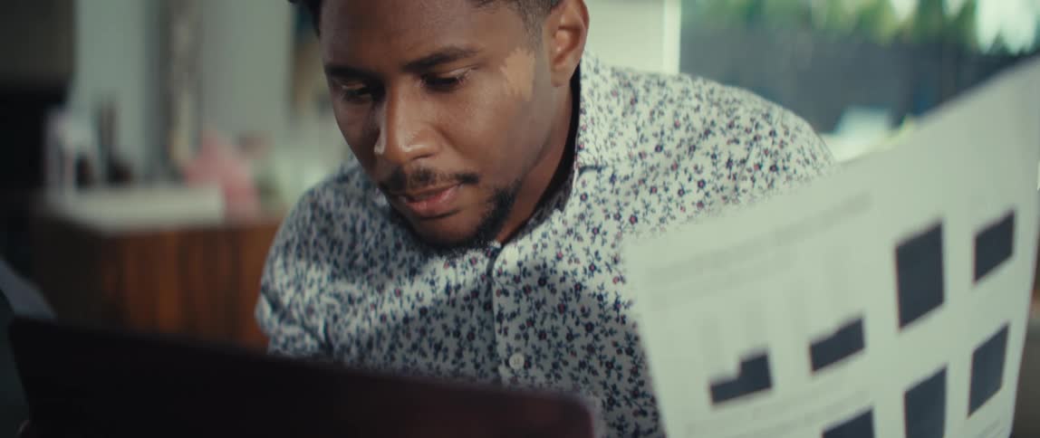 Young adult black man wearing a white dress shirt sitting down and reading from pieces of paper with a contemplative facial expression in a living room indoors during day time