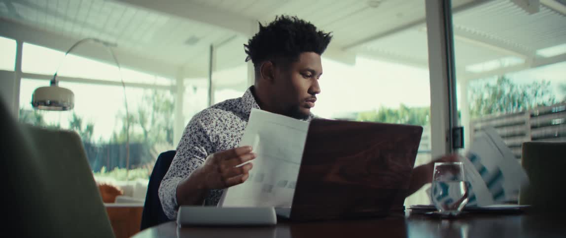 A young black man sits behind a desk inside an office with a laptop in front of him sorting documents on the desk with a glass of water beside him at midday