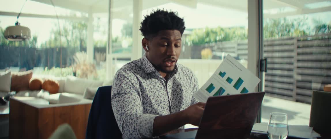 Man sitting at a table holding documents as he has a conversation on a laptop computer