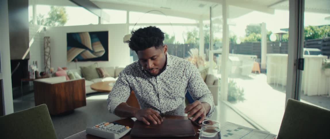 Young adult black man walking into a living room while looking at a smartphone on his hand then sitting down and opening the lid of a laptop computer while checking papers over a wooden table indoors during day time