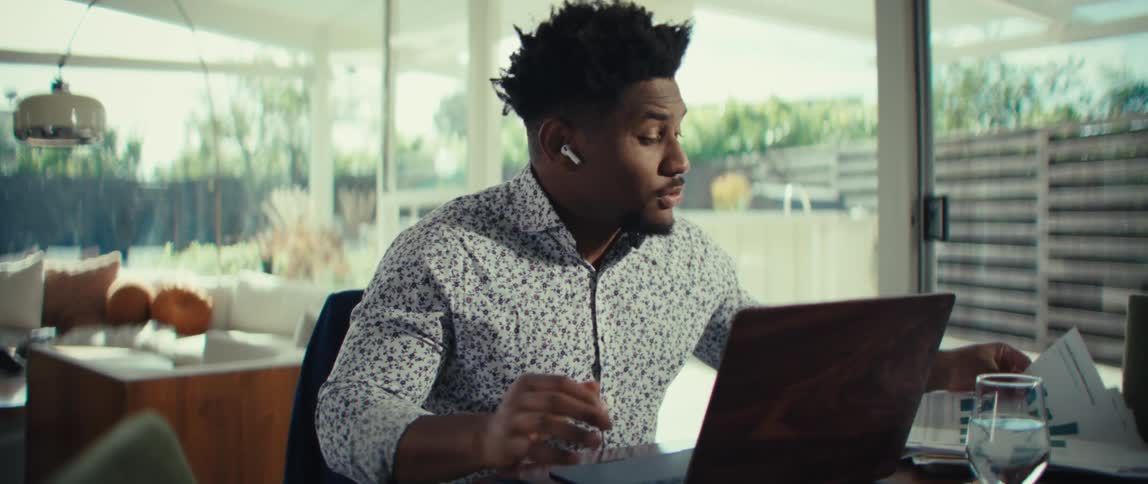 A man wearing a patterned shirt and wireless earbuds sits at a dining table and holds up two documents with colorful charts during a video call on a laptop in a bright modern home