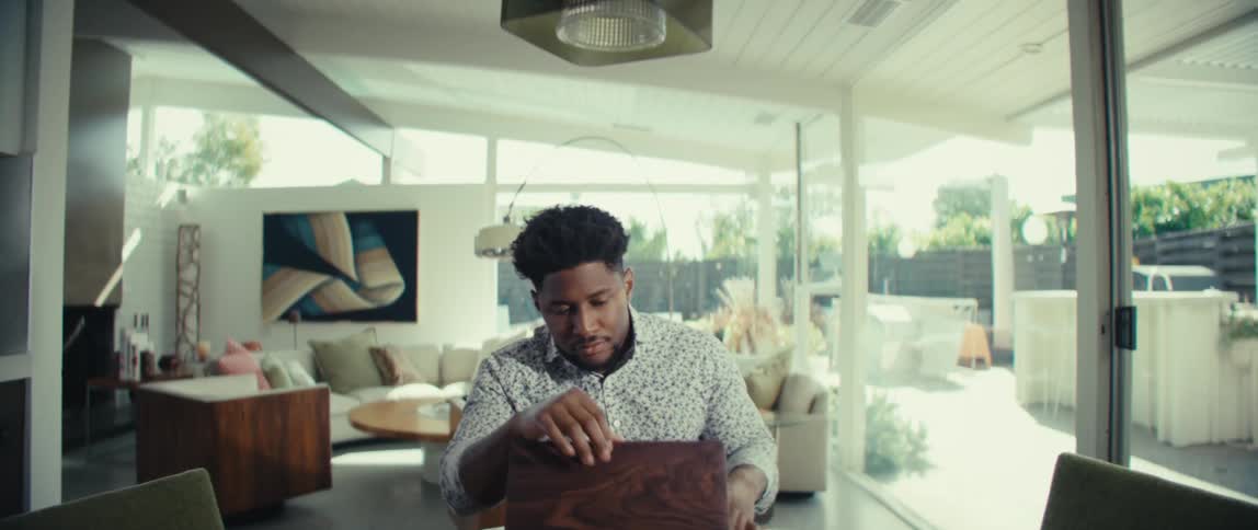 A man with dark hair and a patterned shirt walks into a bright living room while looking at a phone and sits at a table to open a laptop computer