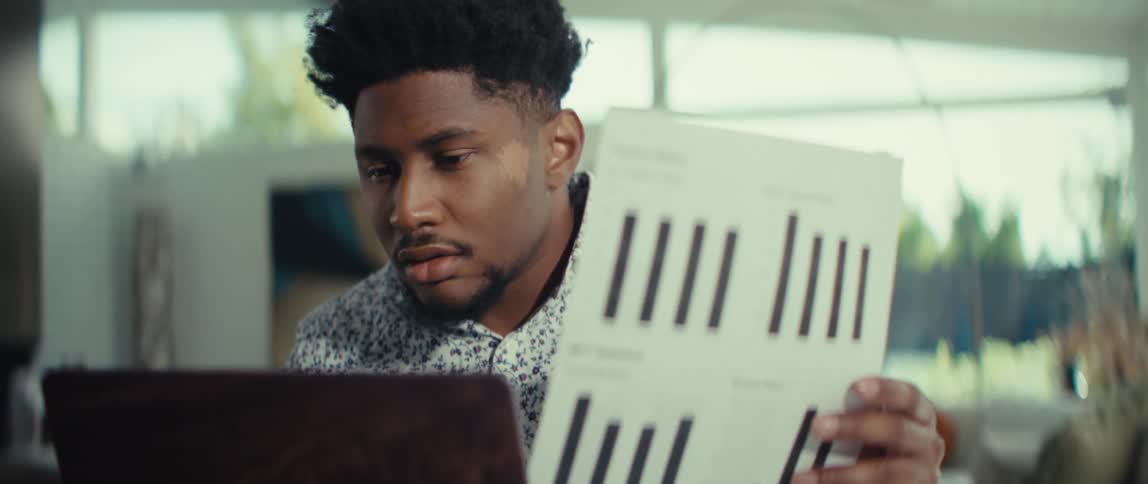 A young man with a focused expression holds a white paper featuring black bar charts and financial text while sitting at a desk with a laptop as the camera shifts focus between the document and his face