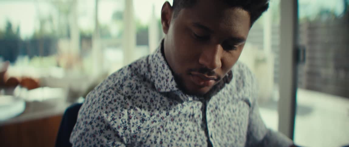 A man in a floral shirt sits at a desk and uses a white calculator while a laptop stays open nearby