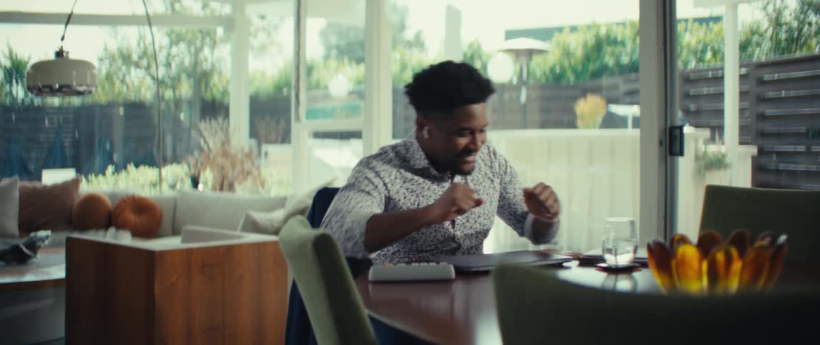 A man with dark hair and a patterned shirt sits at a table in a bright room and reacts with excitement and laughter while closing a laptop computer and enjoying a glass of water