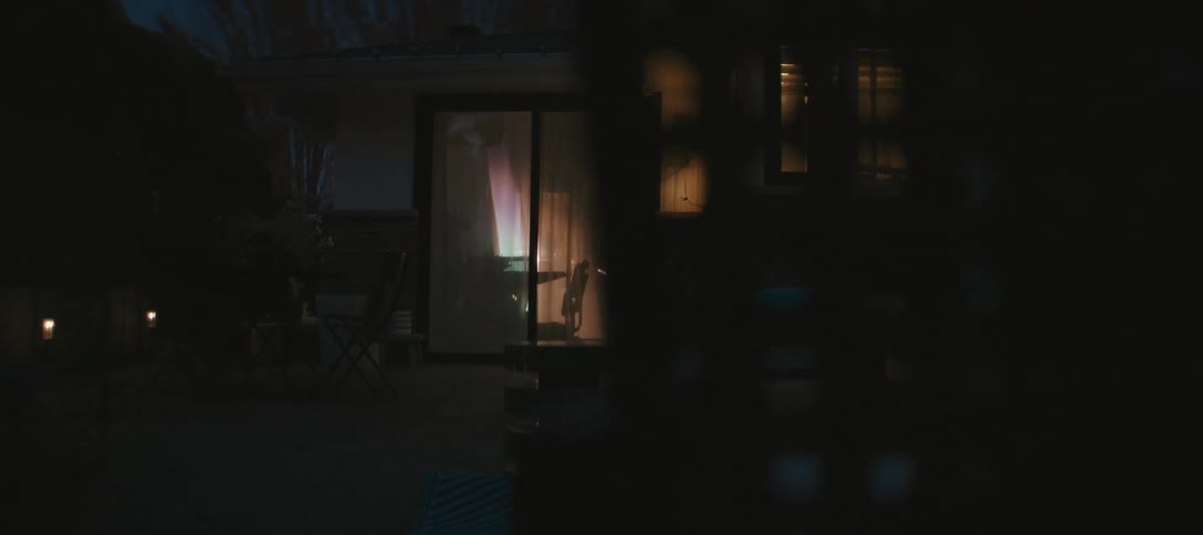 A dimly lit backyard at night reveals a house with warm light glowing through a sliding glass door