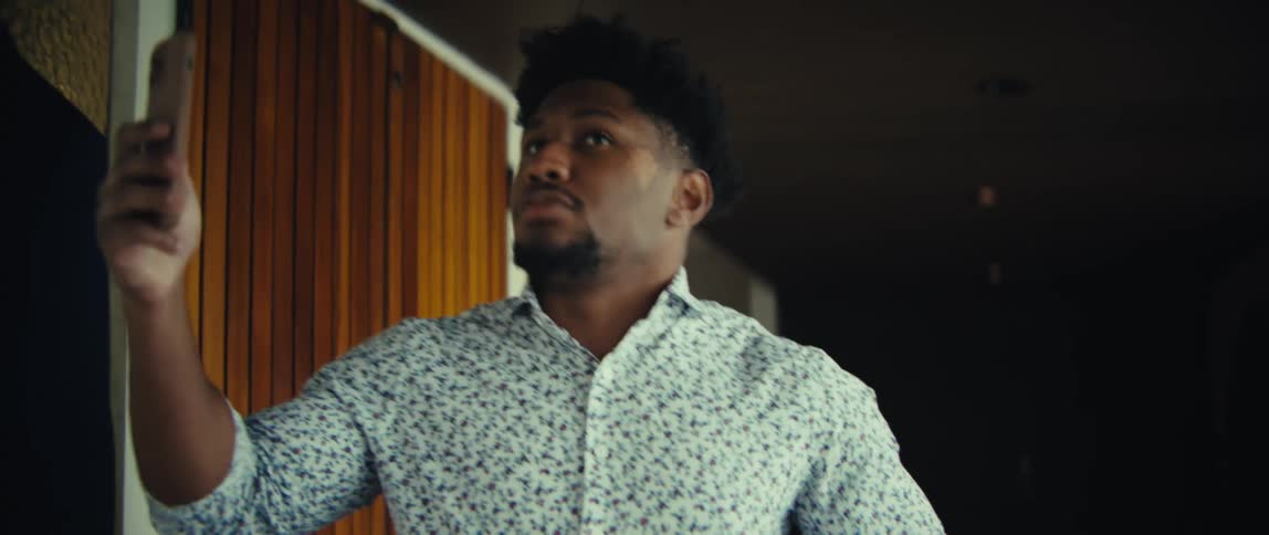 The man is holding his phone up and looking at the screen as if he is on a video callhe appears to be talking and gesturing with his free hand while standing in a room with woodpaneled walls