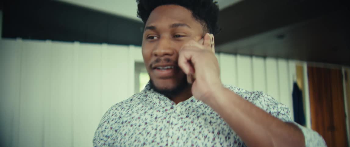 A man with short dark hair and a beard is smiling while talking on a mobile phonehe is wearing a lightcolored patterned shirt and stands in a modern brightly lit indoor space
