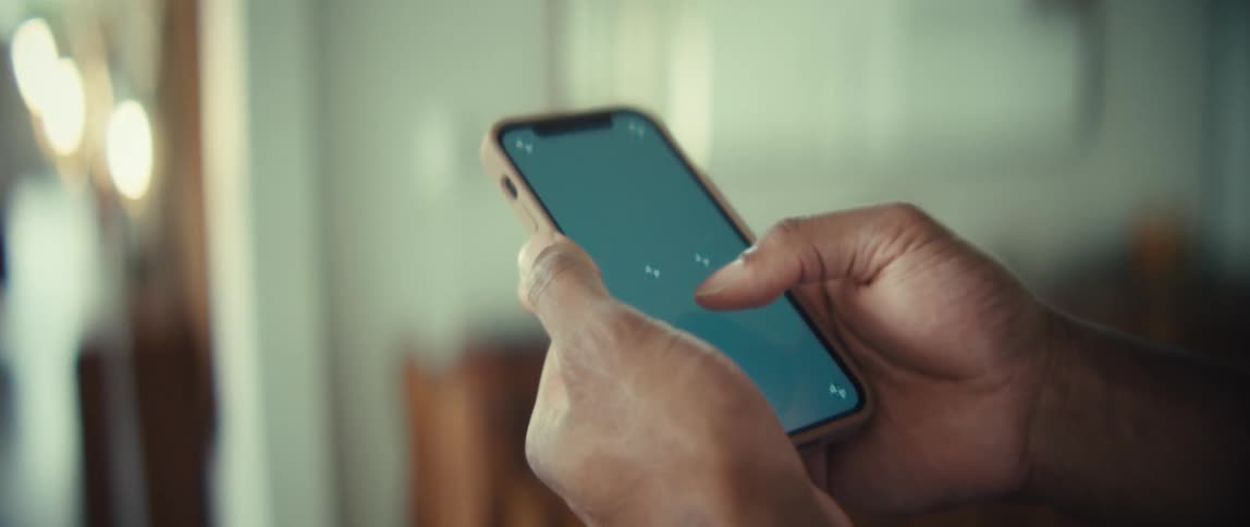 Man stands in a room holding a smart phone with both hands as he moves his thumb across the screen