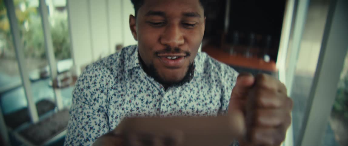 A man wearing a floral shirt looks at a phone and speaks with an animated expression before raising his arms and laughing in a bright indoor setting