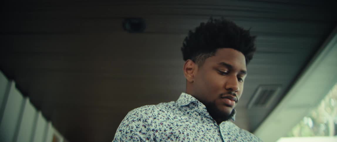 A man wearing a patterned shirt walks through a bright modern house while looking at his phone with a serious expression