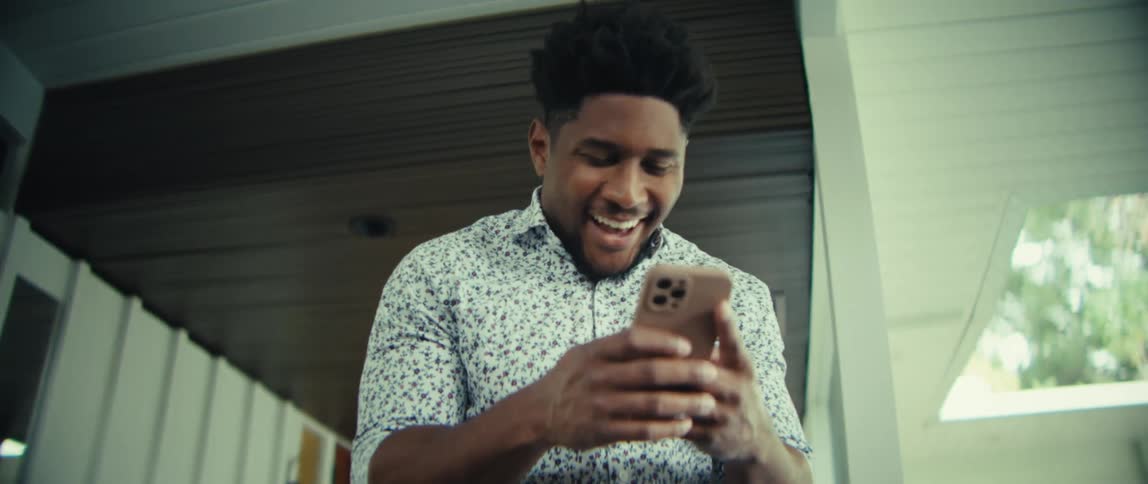 A man wearing a patterned shirt and blue trousers stands on a bright modern house while looking at a phone and laughing with an excited expression