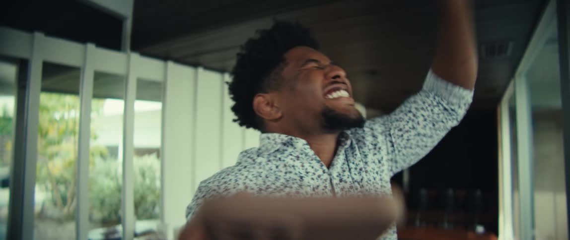 A man wearing a floral shirt looks at a phone and laughs with a wide smile before raising a fist in a celebratory gesture in a bright indoor setting