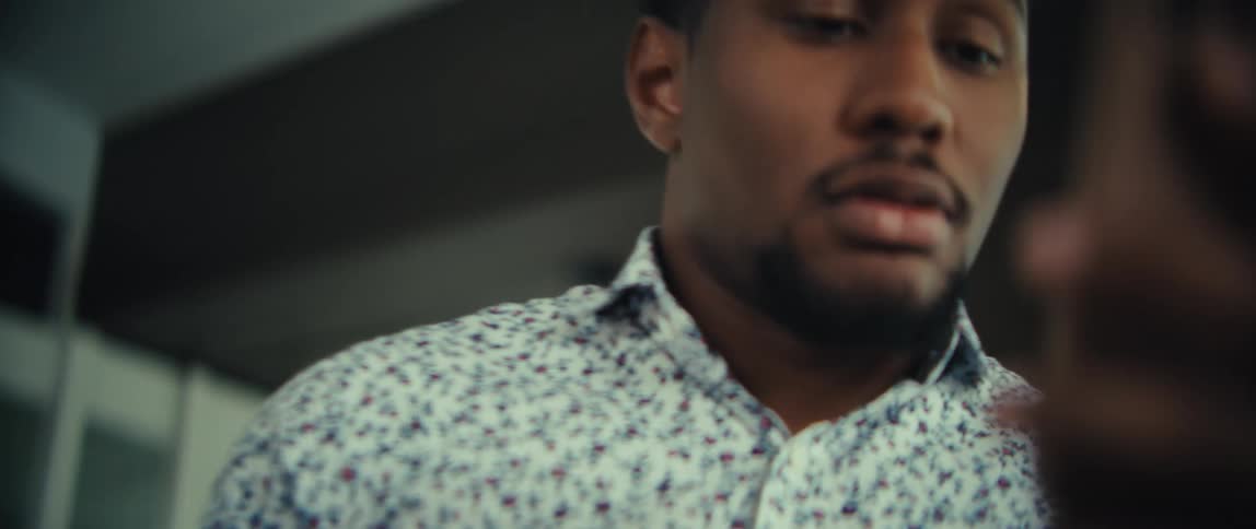 A man with a beard and a patterned shirt is looking down at his smartphone the camera zooms in close to his face as he focuses on the screen with a serious expression