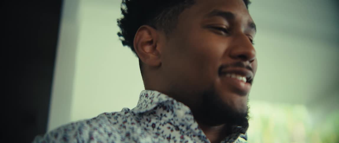 A young man with a short beard and an afro hairstyle looks at his smartphone with a neutral expressionhe then breaks into a warm smile and closes his eyes as the camera moves in for a closeup on the side of his face