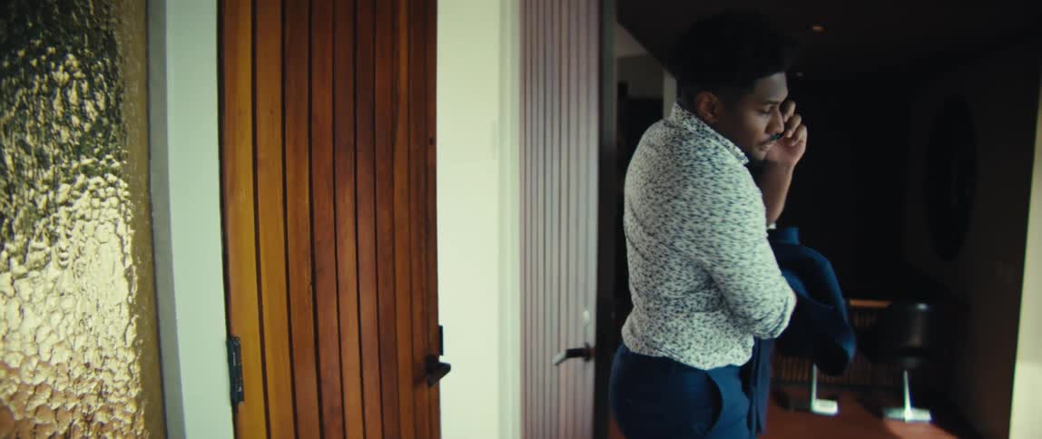 A man wearing a patterned shirt and blue trousers enters a house while talking on a phone and carrying a suit jacket before leaning against a wooden door