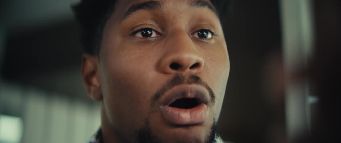 A black man looks at his smartphone and reacts with extreme surprise opening his mouth wide and shouting in excitement while wearing a blue patterned shirt in a brightly lit indoor setting