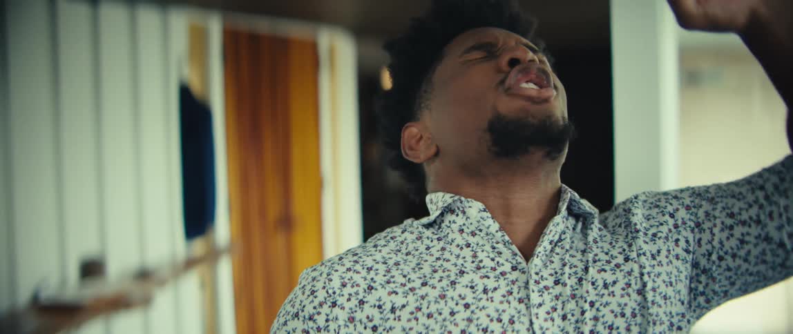 A man wearing a floral patterned shirt and blue trousers speaks on a mobile phone and laughs with a wide smile before pumping his fist in a celebratory gesture in a bright indoor setting