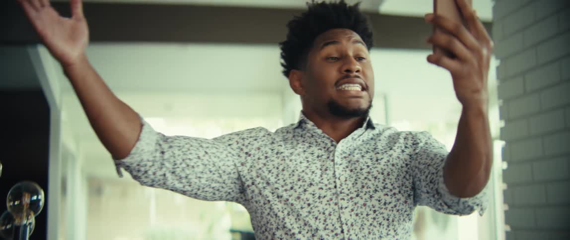 A man wearing a floral shirt looks at a phone and speaks with an animated expression before raising his arms and laughing in a bright indoor setting
