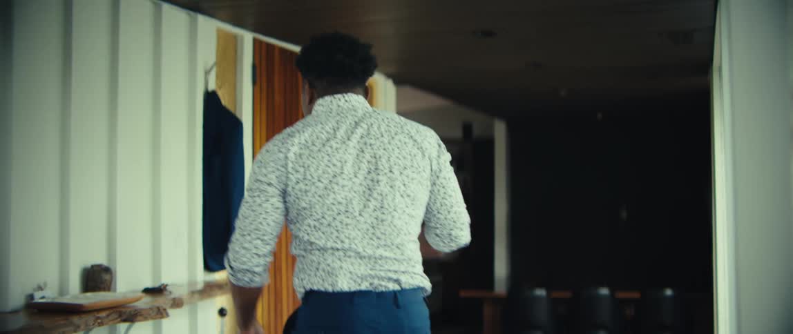 A man in a patterned shirt walks through a modern home while talking on his mobile phone he stops to grab a dark blue jacket hanging on the wall before exiting through a doorway