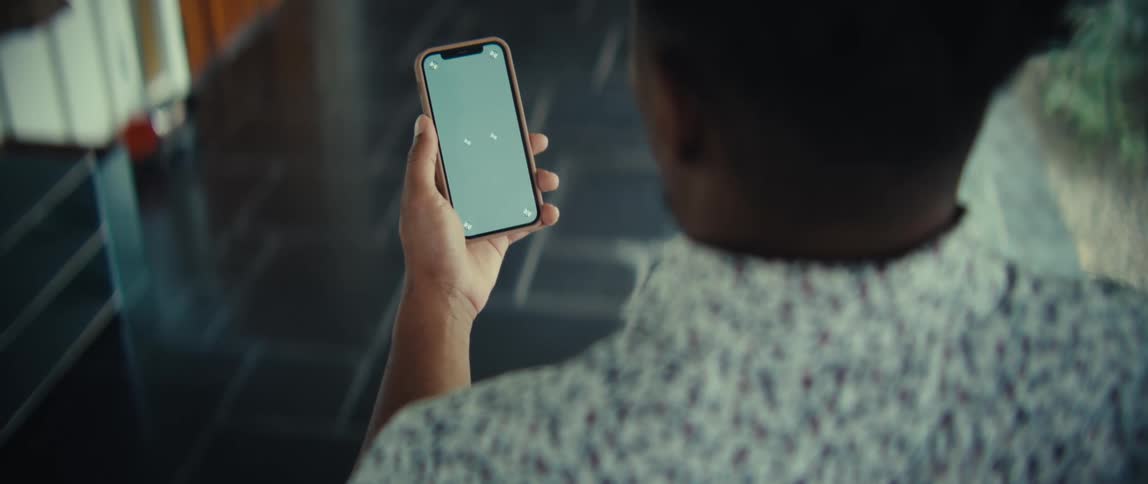 A man in a patterned shirt walks down a hallway while holding a smartphone with a green screen interface the camera follows him from behind capturing his movement across a dark tiled floor toward a bright doorway