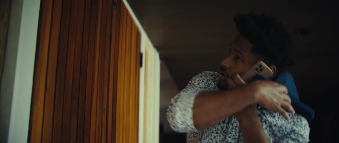 A black man in a patterned shirt talks on his smartphone while simultaneously pulling a blue jacket over his shoulders as he enters a room through a wooden slatted door