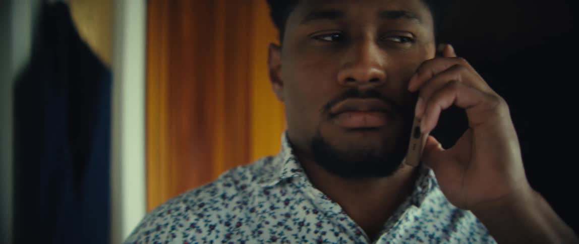A man wearing a floral patterned shirt walks through a house while engaged in a phone conversation his expression shifts from focused listening to a wide joyful laugh as the call continues