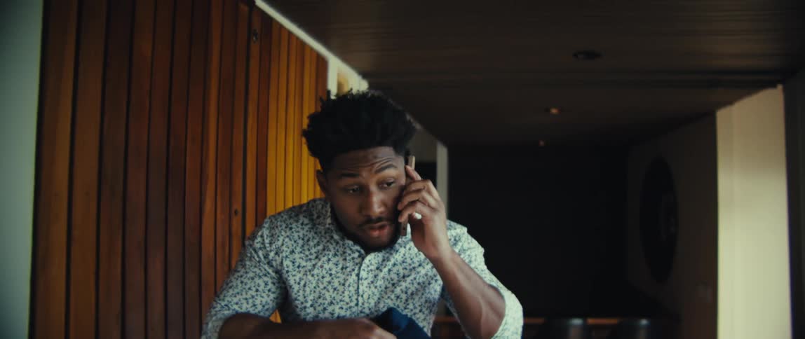 A man wearing a floral shirt and blue trousers walks through a bright modern house while speaking on a mobile phone before hanging a coat on the wall