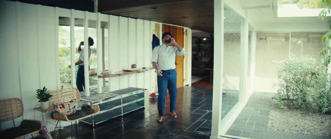 A man hangs a blue jacket on a wall hook and walks through a modern hallway while talking on his phone as he approaches the camera he laughs heartily during his conversation