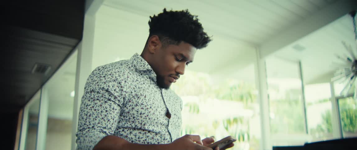 Young adult black man walking indoors while texting and then stopping and smiling with excitement during day time