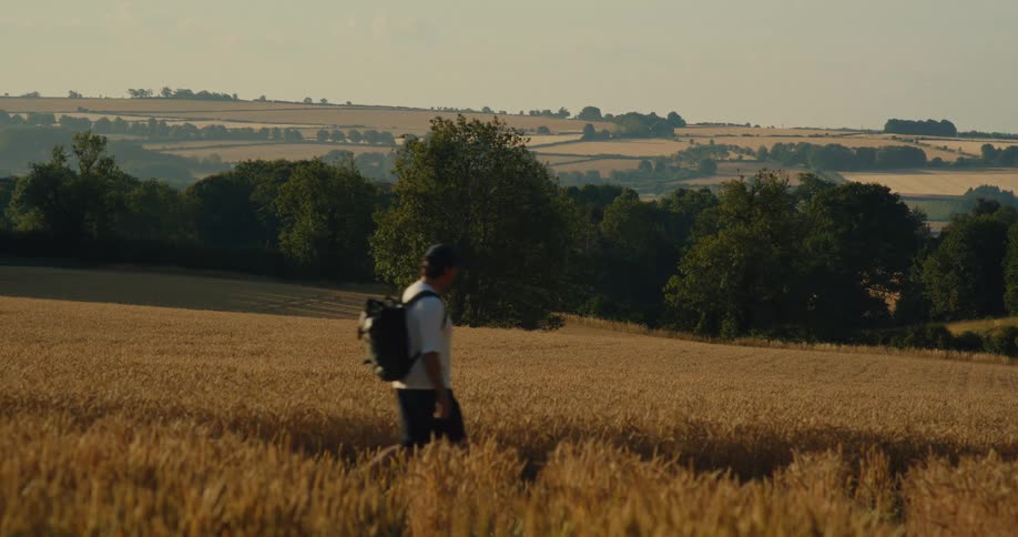 Young man wearing a backpack walks on a path through rural farmland on a hill