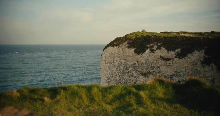 A rugged white chalk cliff stands prominently against a calm blue sea under the soft light of a late afternoon sun in the foreground vibrant green grass sways gently on a nearby ridge framing the vast coastal landscape
