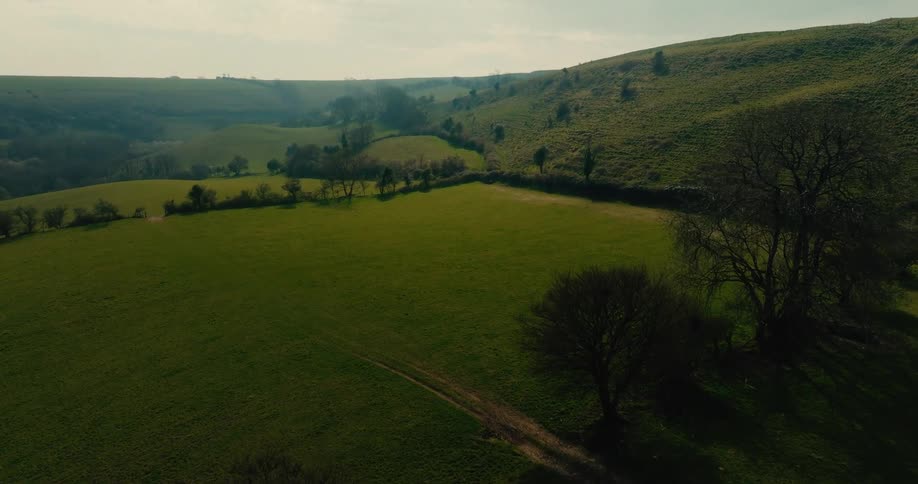 An aerial camera sweeps across a rolling green valley where sunlight highlights the contours of the grassy hills and scattered trees the vast landscape is marked by natural hedgerows and a narrow dirt path that winds through the fields toward a hazy hori