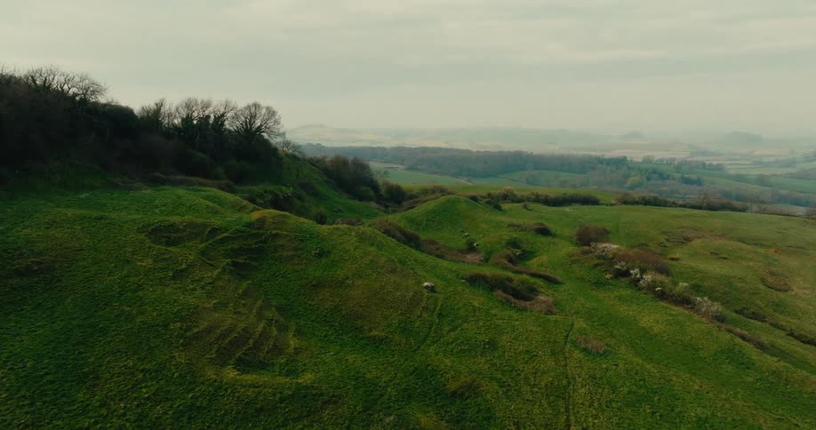 An aerial shot captures a rolling green hillside under a pale overcast sky the camera slowly pans across the textured terrain revealing small white animals grazing on the steep grassy slopes