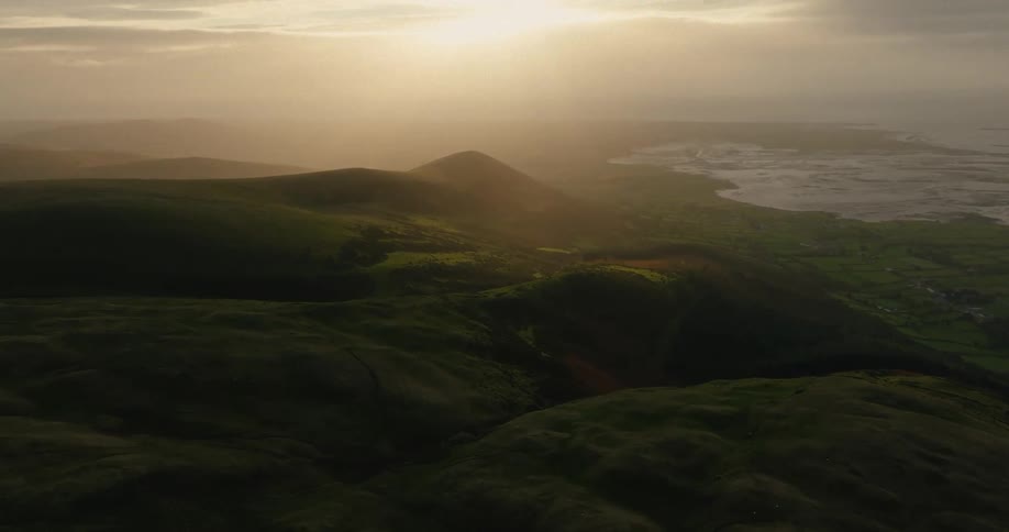 A sweeping mountain range at sunset where the golden light of the sun breaks through a hazy overcast skyin the background a calm body of water meets a rugged coastline creating a peaceful contrast between the dark rolling hills and the glowing hor