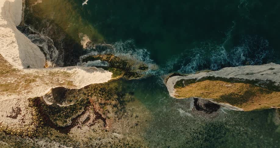 This topdown aerial footage captures turquoise ocean waves crashing against the base of jagged grasstopped coastal cliffs the sunlight casts a warm glow over the landscape highlighting the clear water and rocky seabed below