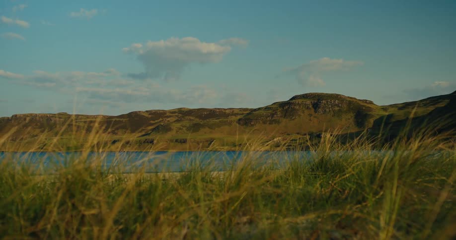 Grass in the foreground frames a view of a lake and hills under a sky with clouds