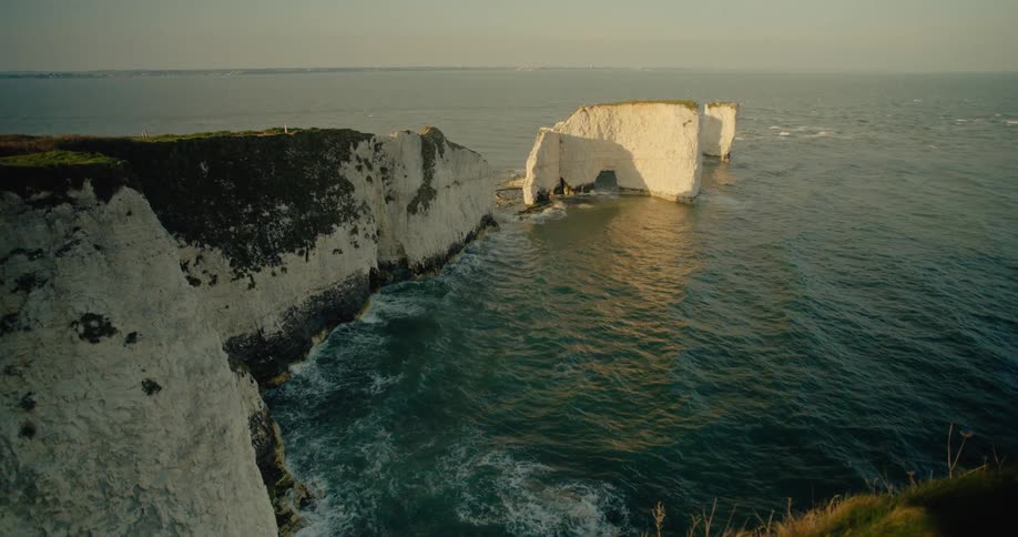 A high angle view shows white chalk cliffs and a natural stone archway in the ocean as waves crash against the base during a clear sunset