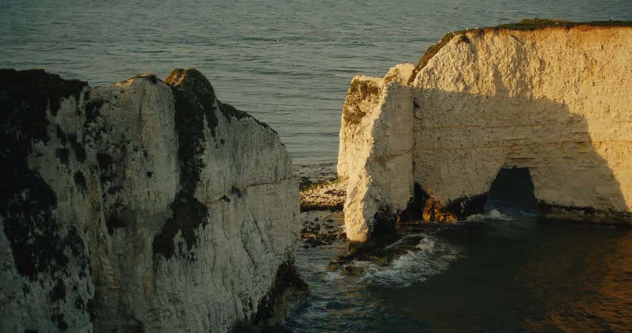 A high angle view shows white chalk cliffs and a natural stone archway in the ocean as waves crash against the base during a clear sunset