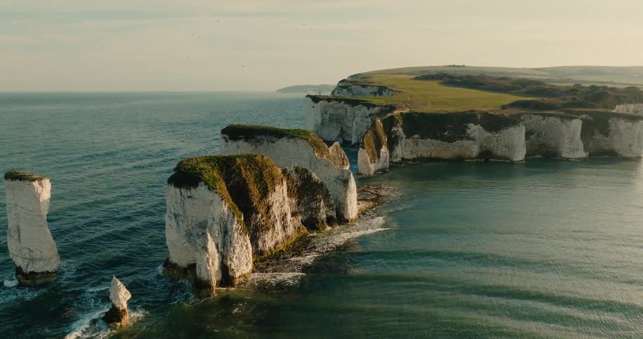 Tall white chalk cliffs and isolated sea stacks stand prominently along a rugged coastline while calm ocean waves crash against the stone base under a clear sky
