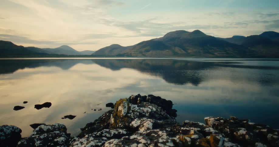 A still lake reflecting a large mountain range under a pale morning sky