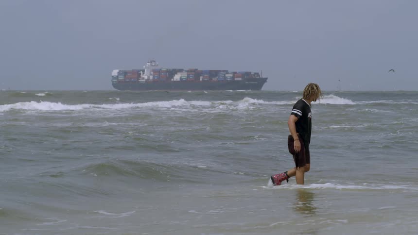 A man wades in the water as a cargo ship tilts in the water
