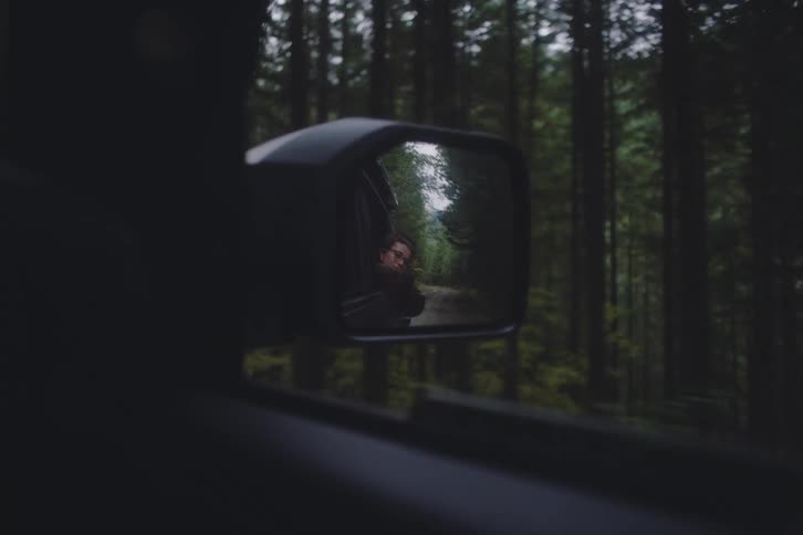 A view through a side mirror shows a woman leaning out of a car window and smiling while driving through a dark evergreen forest on a cloudy day