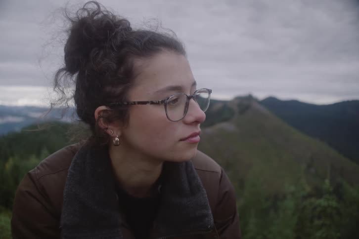A close view shows a woman with glasses and dark hair in a bun looking out over a vast mountain range under an overcast sky during a cold day
