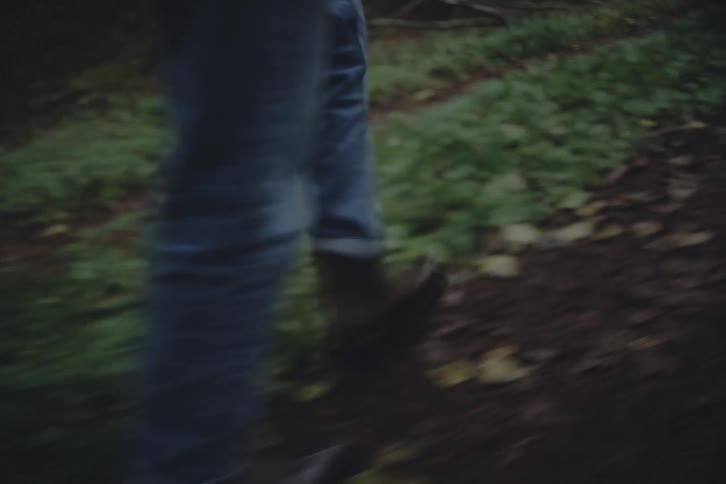 A person wearing blue jeans and brown boots walks along a dark forest path covered in green moss and fallen leaves