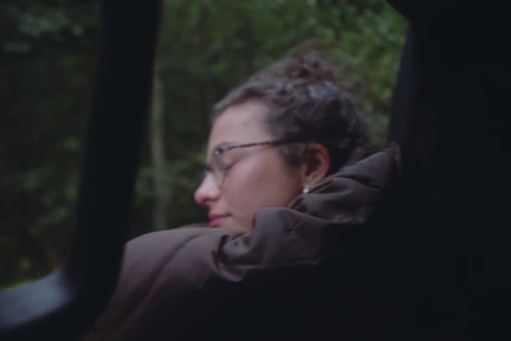Young female wearing glasses leaning over the window of a vehicle driving next to a forest area during day time