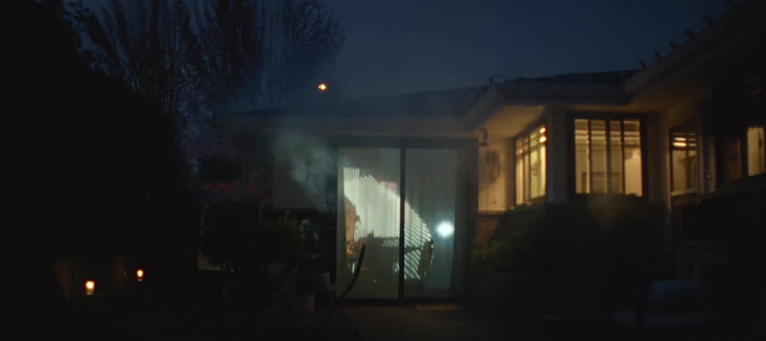 In a smoky outdoor setting at night a projector displays shifting fanlike geometric patterns onto the glass doors of a housethe camera slowly zooms out revealing more of the dark patio and the illuminated windows of the home as the lig