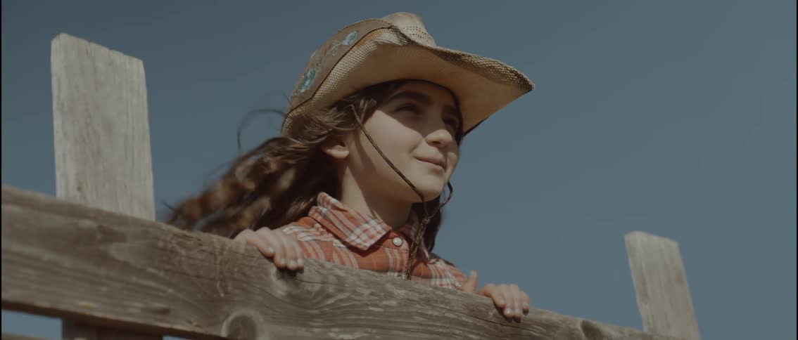 Young teenager girl wearing a plaid shirt and cowboy hat standing next to a wooden fence and smiling outdoors during day time