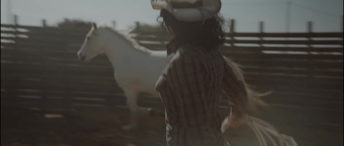 A woman in a cowboy hat and plaid shirt practicing with a lasso in a dusty corralin the background a white horse trots along the wooden fence as the woman swings her rope in a circular motion